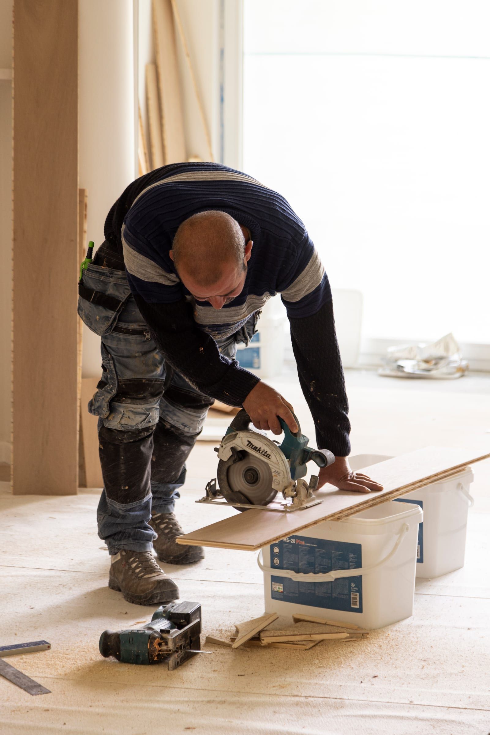 Découpe d’une lame de parquet en chêne à la scie circulaire, devant une colonne du salon en cours de finition