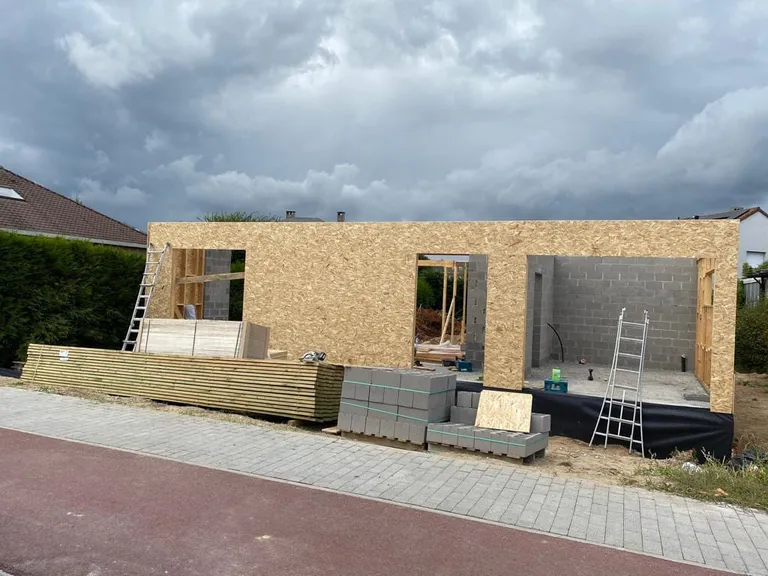 Construction en ossature bois et panneaux sur fondations en blocs, vue depuis la rue, bois empilé devant