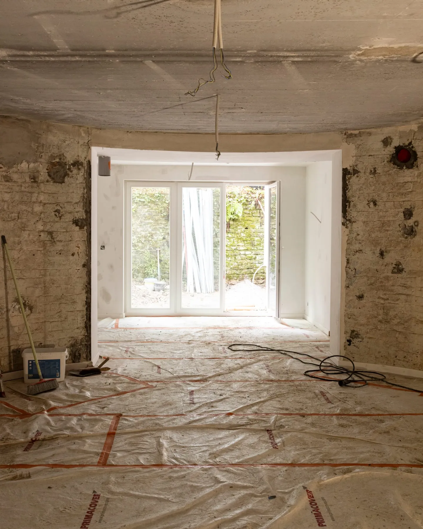 Pièce mise à nu dans une maison en rénovation : murs en brique, plafond en béton, nouvelle baie ouverte sur le jardin